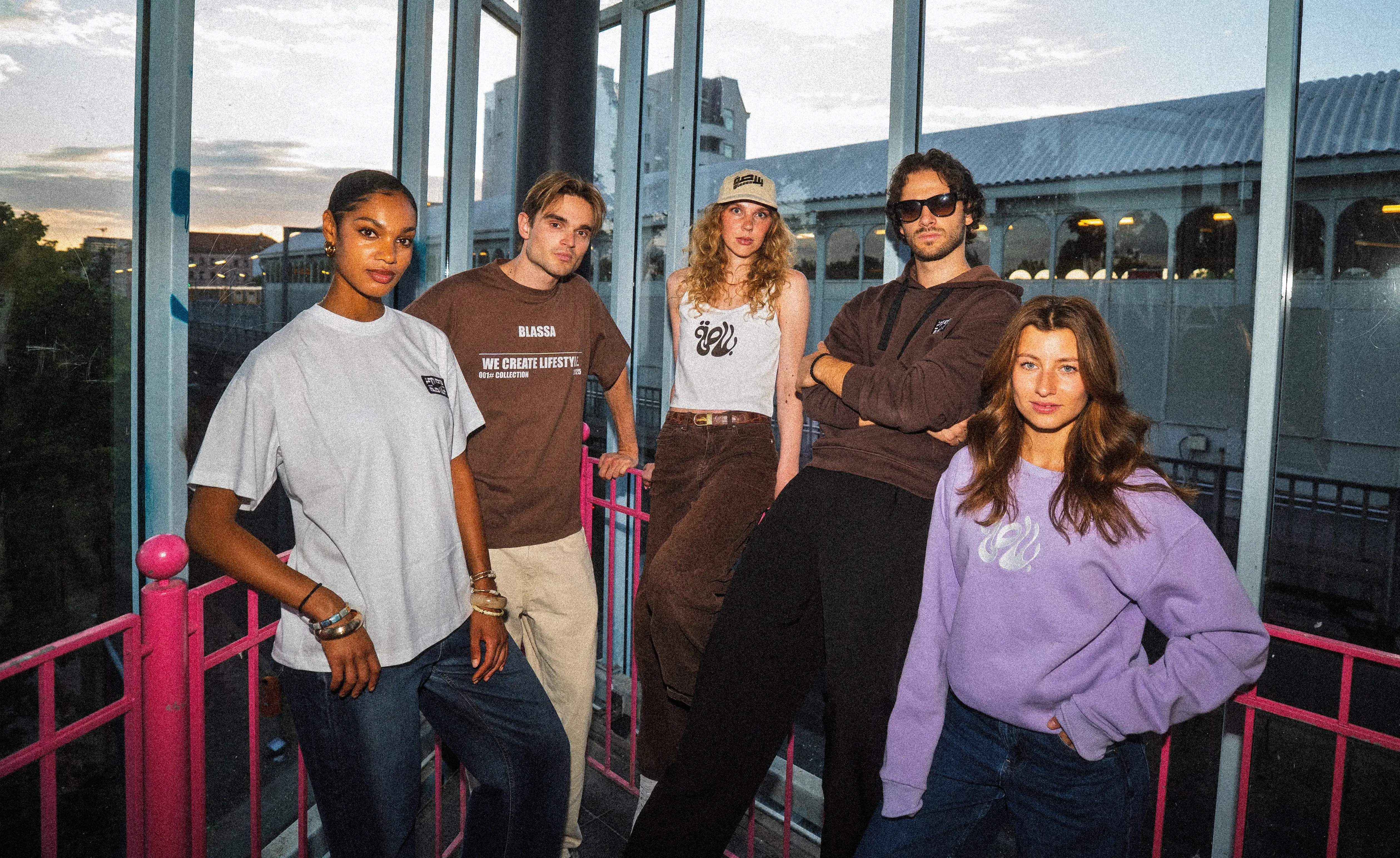 Five friends in Blassa tees, a tank top, a hoodie and a lilac sweatshirt on a pink-railed footbridge at dusk